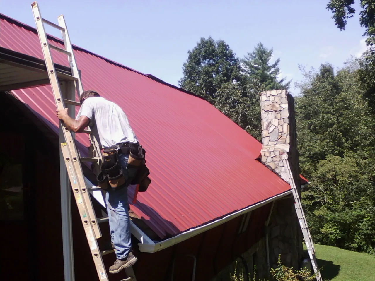 Roofer on a ladder beside a red metal roof and stone chimney — Soffit & Fascia Repair by Roofing Brothers in Lewistown